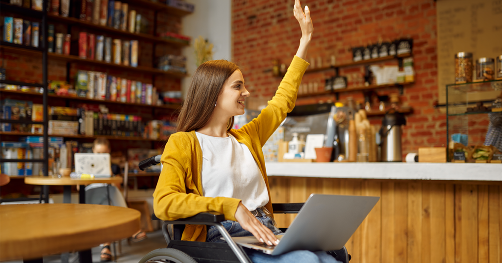 Eine junge Frau sitzt in einem Rollstuhl in einem Café und hat einen Laptop auf ihrem Schoß. Sie lächelt und hat den linken Arm zur Begrüßung erhoben. Eine barrierefreie Website ist für sie und viele andere Menschen sehr wichtig.
