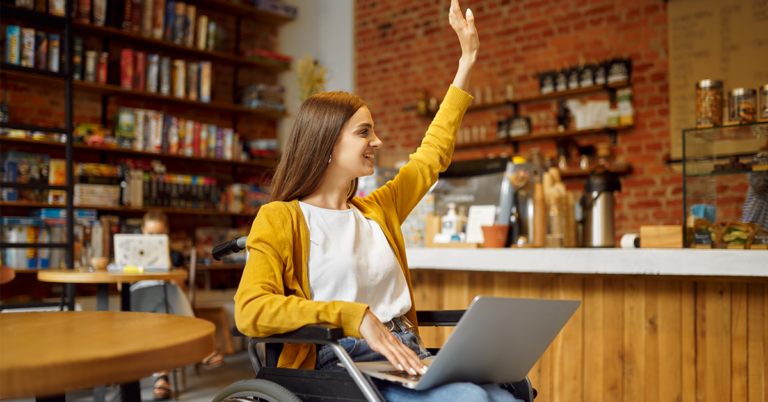 Eine junge Frau sitzt in einem Rollstuhl in einem Café und hat einen Laptop auf ihrem Schoß. Sie lächelt und hat den linken Arm zur Begrüßung erhoben. Eine barrierefreie Website ist für sie und viele andere Menschen sehr wichtig.