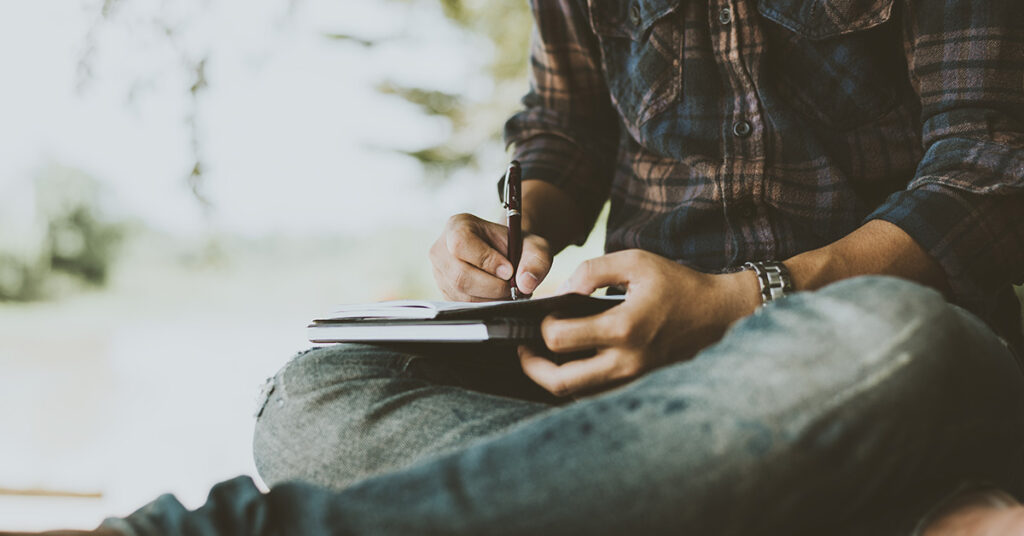 Beitragsbild zum Thema Storytelling: Eine Person in Jeans und karierte Hemd sitzt im Schneidersitz und schreibt in ein Notizbuch.