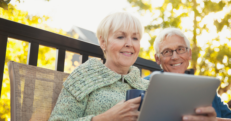 Das Bild zeigt ein Seniorenpächen, das auf einer Bank im Freien sitzt. Sie hält ein Tablet in der Hand und schaut lächelnd darauf, er sitzt neben ihr und schaut sie lächelnd an.