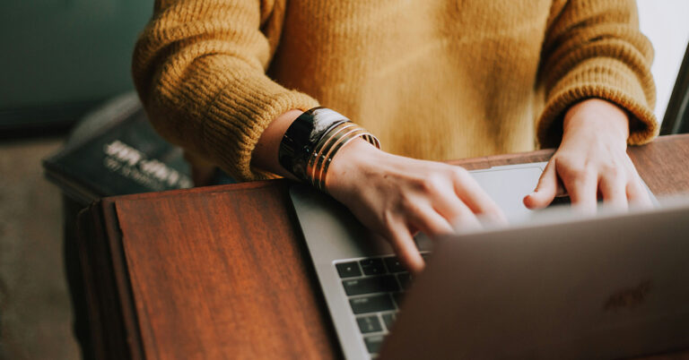Eine weibliche Person im senfgelben Pullover sitzt vor einem Laptop und tippt etwas - eine Szene, die typisch für Online-Weiterbildungen ist.